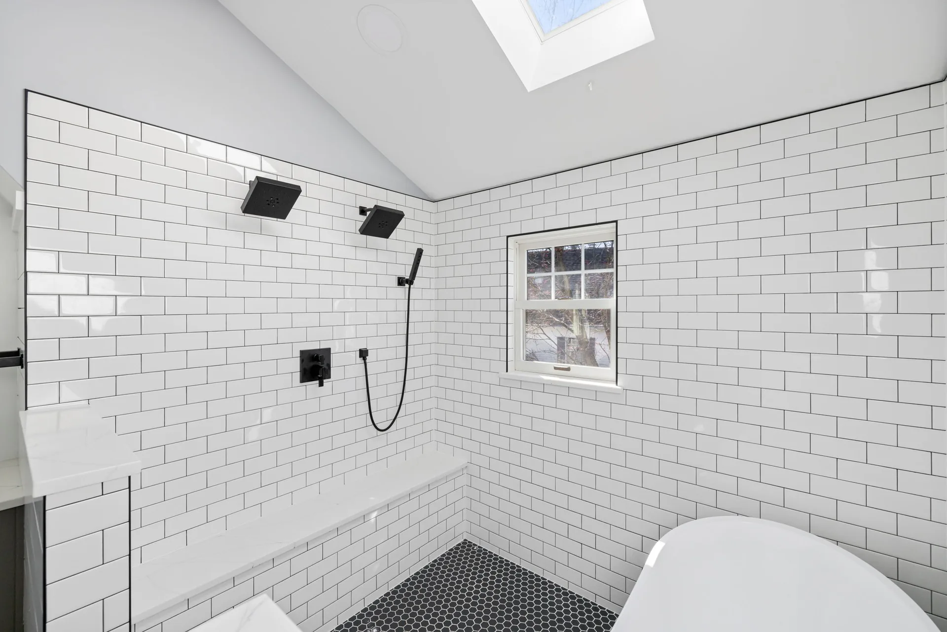 Finished bathroom with white tile shower, black fixtures, freestanding tub, and skylight.