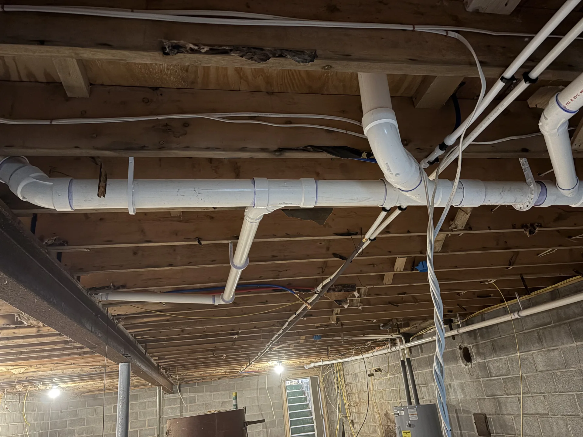Overhead PVC drain piping installed between open basement floor joists.